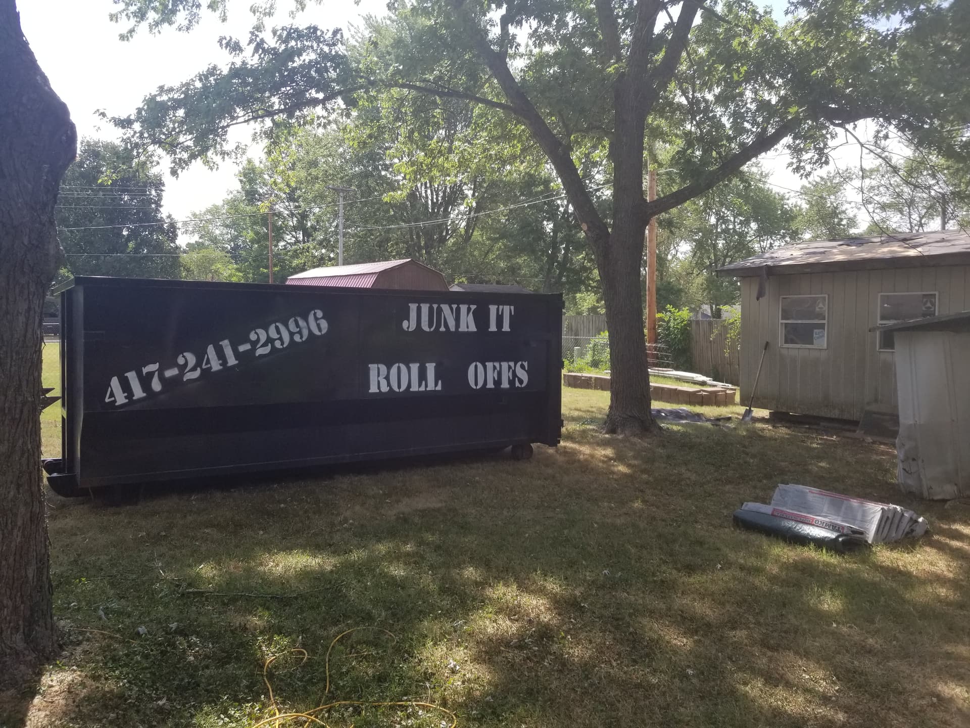 Black 25-yard roll-off dumpster from Junk It Roll Offs placed in a residential backyard in Fordland, MO, with roofing shingles nearby.